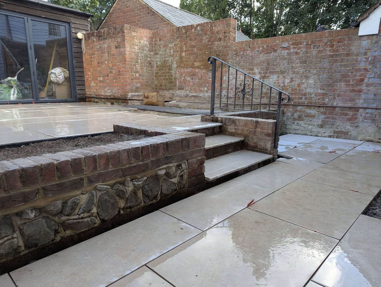 Rebuilt walled garden: brick and flint retaining wall, stone steps with wrought iron rail, and rain-washed porcelain paving